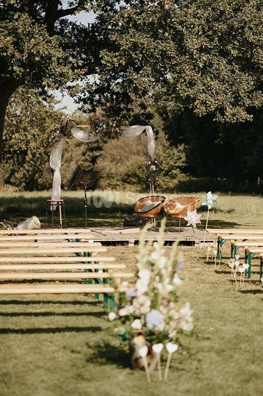 Chaises et arche décorée pour une cérémonie en plein air, entourées de bancs en bois et de verdure.