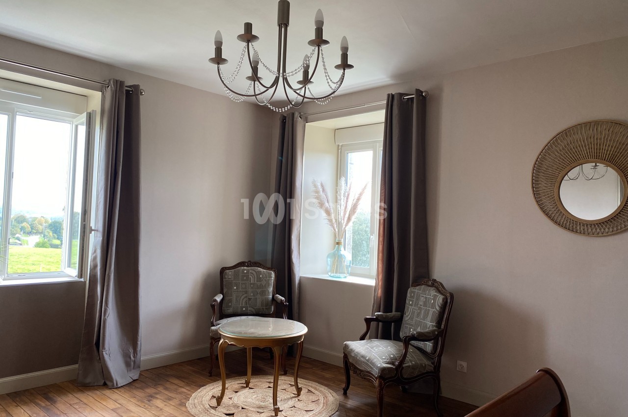 Deux fauteuils et une table ronde sur un tapis, près de deux fenêtres avec rideaux, dans une pièce lumineuse.