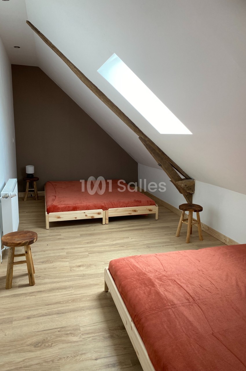 Deux lits avec draps orange dans une chambre mansardée lumineuse, parquet en bois clair et poutre apparente.