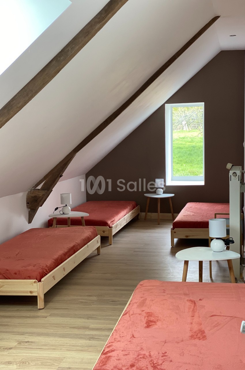 Quatre lits simples avec draps rouges dans une chambre mansardée lumineuse, avec murs blancs et une fenêtre donnant sur un…