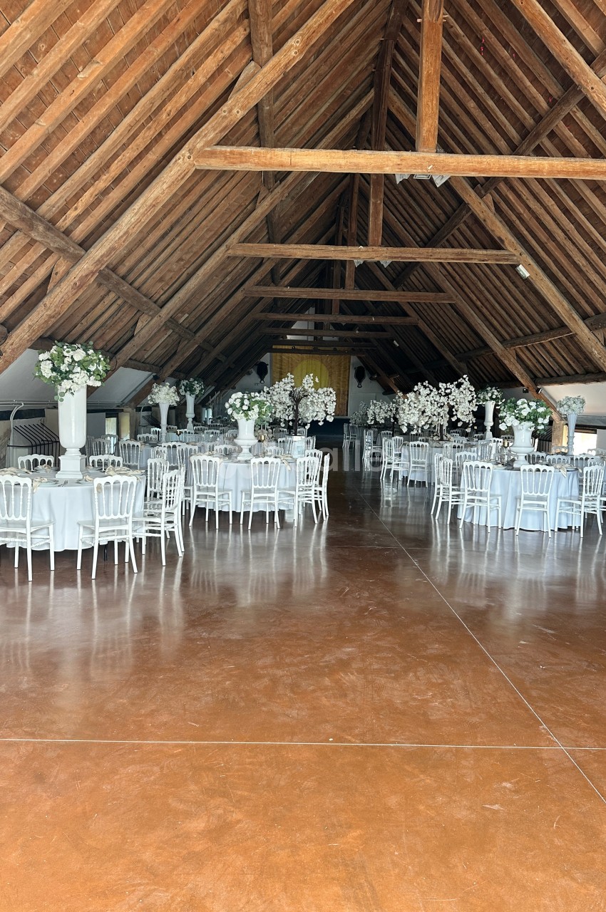 Salle de réception avec charpente en bois, tables rondes décorées de nappes blanches et arrangements floraux.