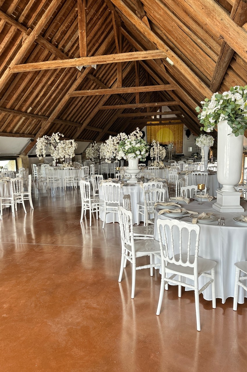 Salle de réception avec tables rondes dressées, chaises blanches et décorations florales sous une charpente en bois.