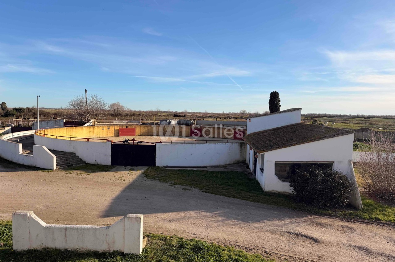 Arène circulaire en plein air entourée de bâtiments blancs, située dans un paysage rural sous un ciel dégagé.