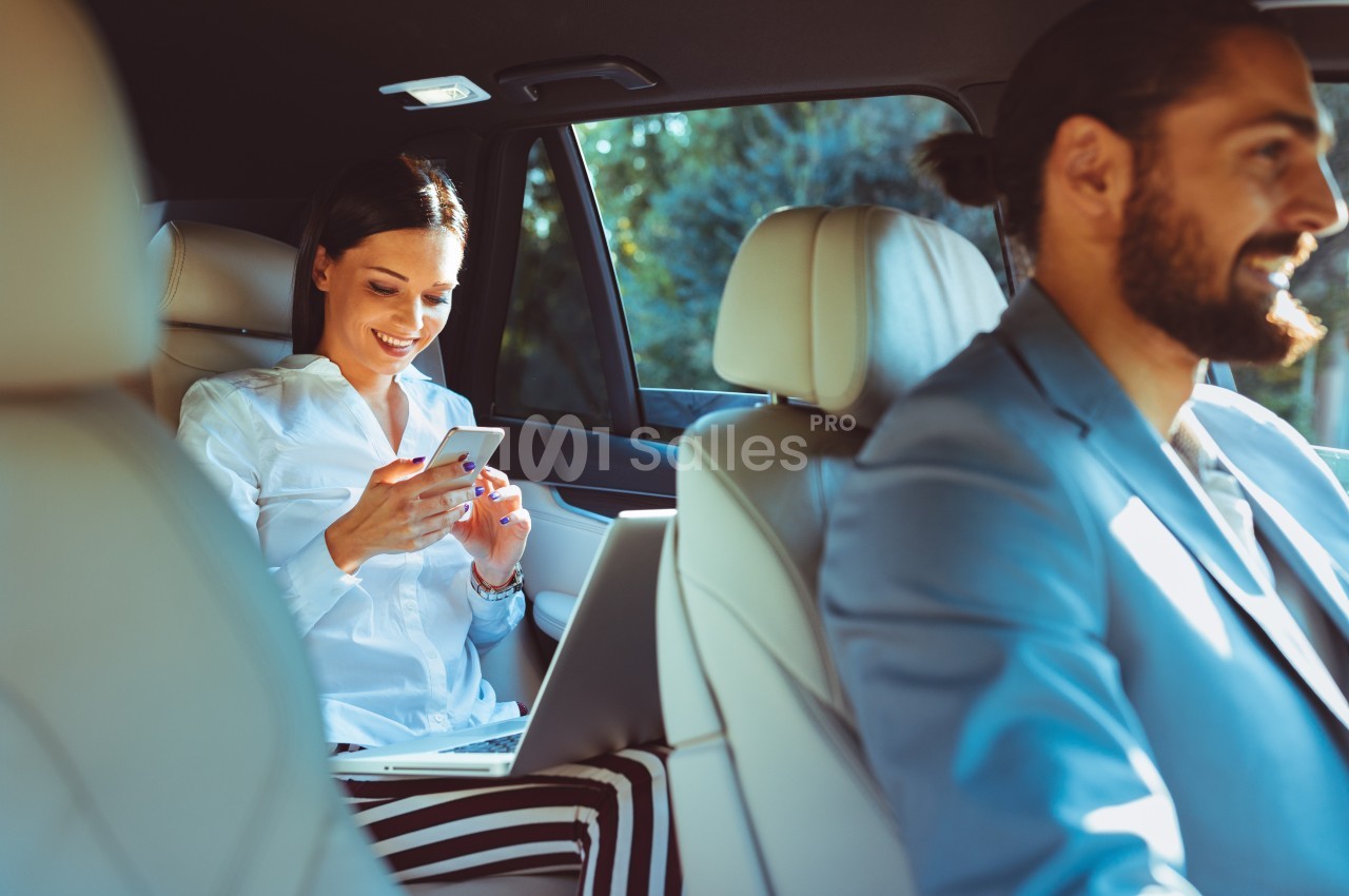 Une femme souriante utilise son téléphone dans une voiture, assise à l'arrière, avec un homme conduisant à l'avant.