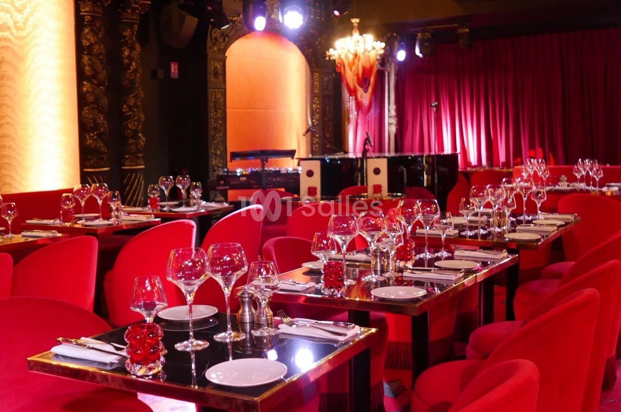 Salle de restaurant élégante avec des tables dressées, des chaises rouges et une scène éclairée en arrière-plan.