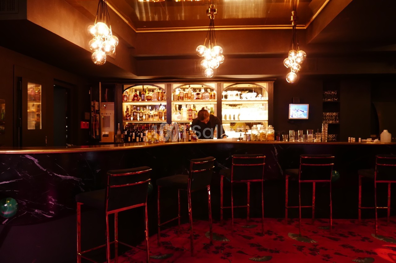 Bar élégant avec comptoir en marbre noir, étagères éclairées remplies de bouteilles et tabourets alignés sur un tapis rouge.