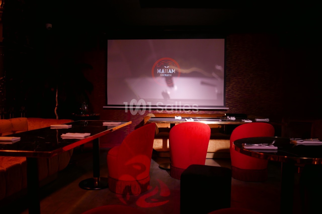 Salle sombre avec des tables, des chaises rouges et un écran de projection affichant un logo lumineux.