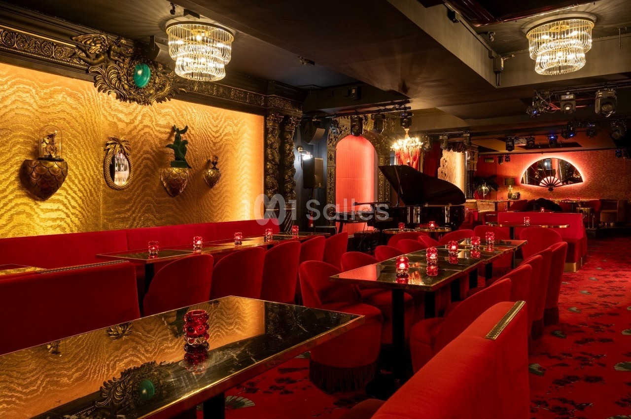 Salle élégante avec des tables et chaises rouges, décor doré, lustres, piano à queue et ambiance tamisée.