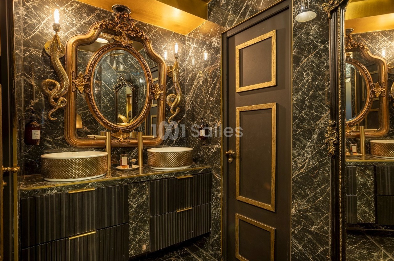 Salle de bain luxueuse avec murs en marbre noir, miroirs dorés ornés de motifs serpentins et lavabos design.