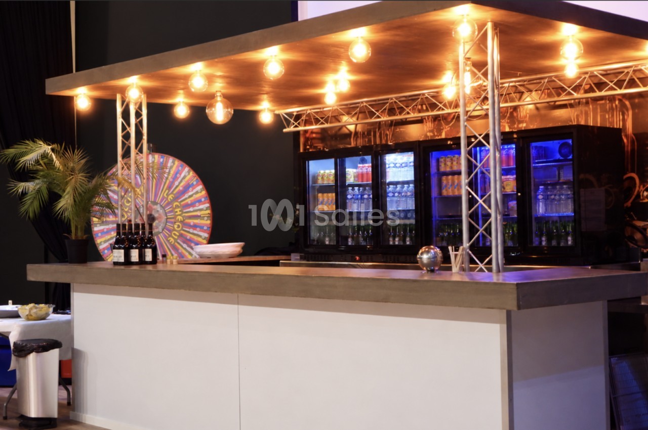 Bar comptoir blanc avec toile de fond colorée, palmier, ampoules suspendues et réfrigérateurs éclairés en arrière-plan.