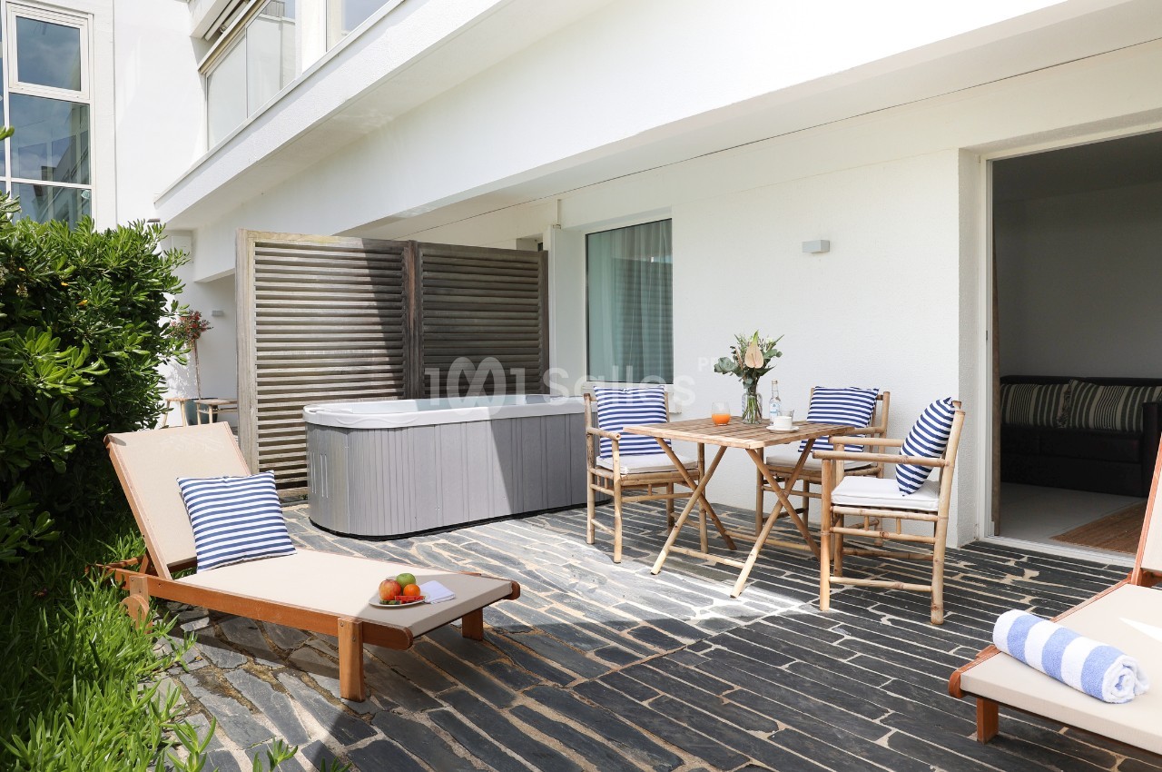 Terrasse avec bain à remous, chaises longues, table en bois dressée et coussins rayés, entourée de verdure.