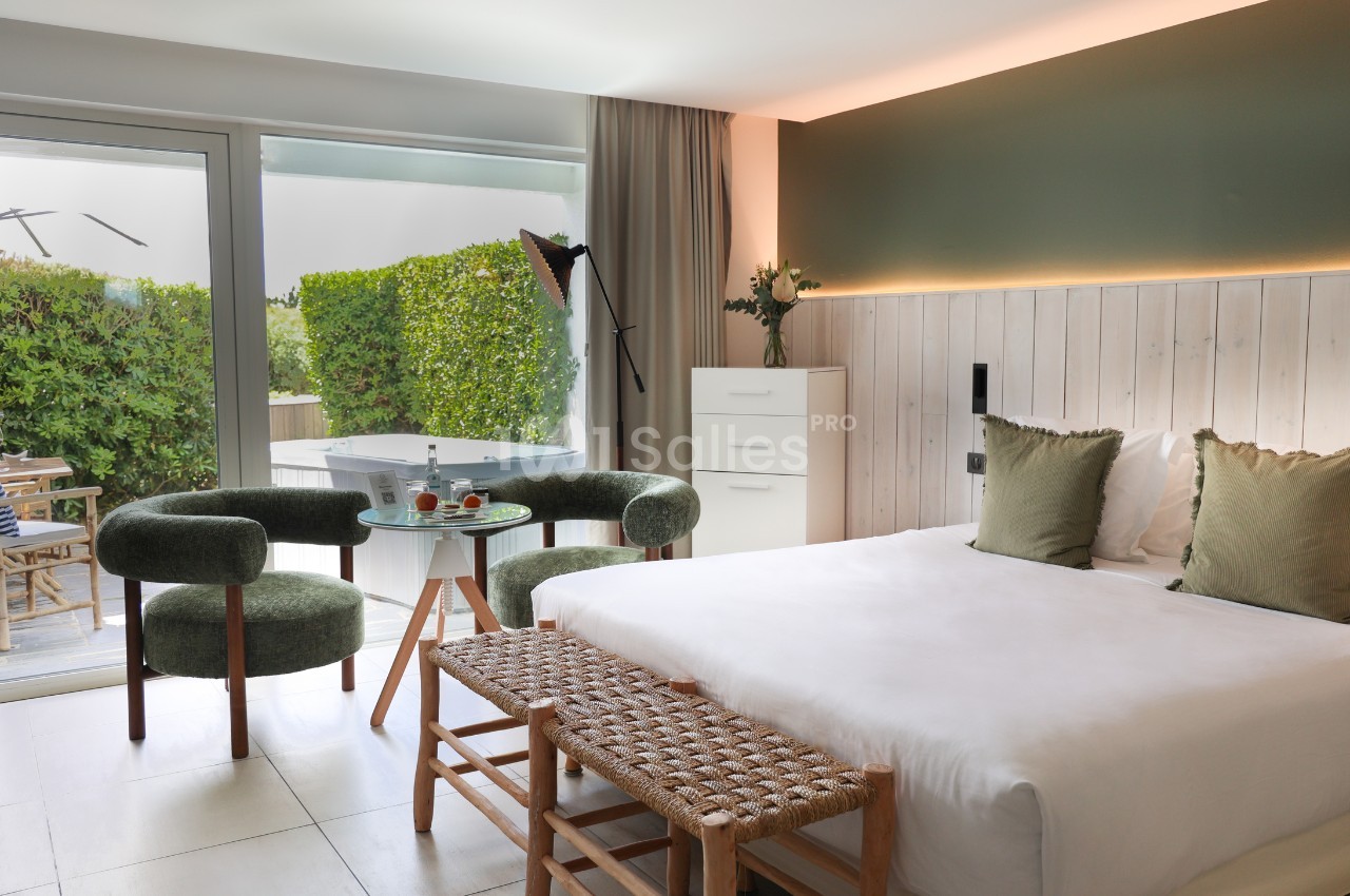 Chambre lumineuse avec lit double, fauteuils verts, table basse, et accès à une terrasse avec jacuzzi et végétation.