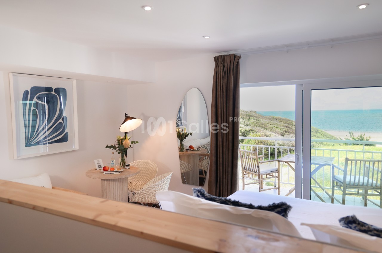 Chambre lumineuse avec lit, table, fauteuil, balcon donnant sur la mer et décoration moderne.