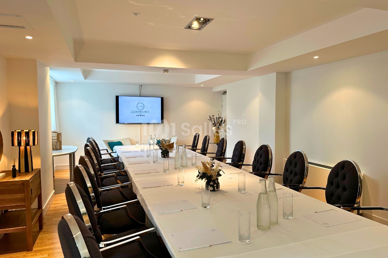 Salle de réunion lumineuse avec une grande table rectangulaire, chaises noires, écran mural et décoration sobre.