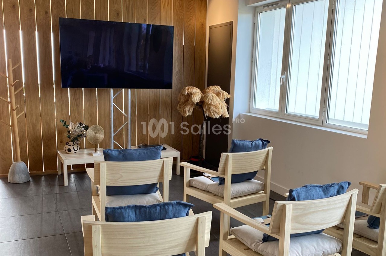 Salle lumineuse avec des chaises en bois, coussins bleus, écran mural et décoration naturelle en arrière-plan.