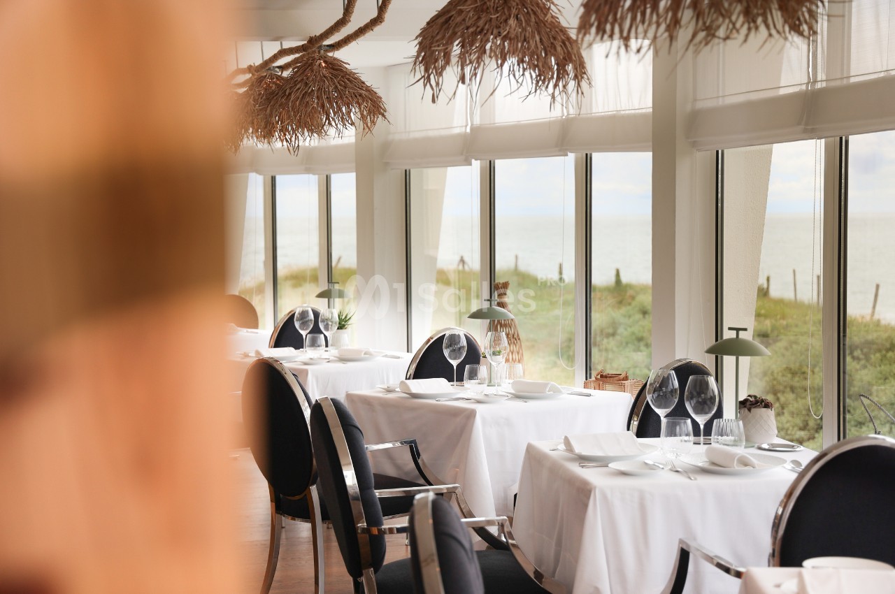 Salle de restaurant lumineuse avec vue sur la mer, tables dressées avec nappes blanches et décoration naturelle.