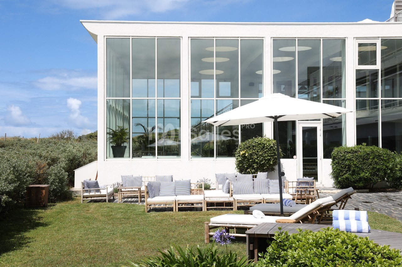 Terrasse aménagée avec mobilier en bois, parasol blanc et plantes, devant une maison aux grandes baies vitrées.