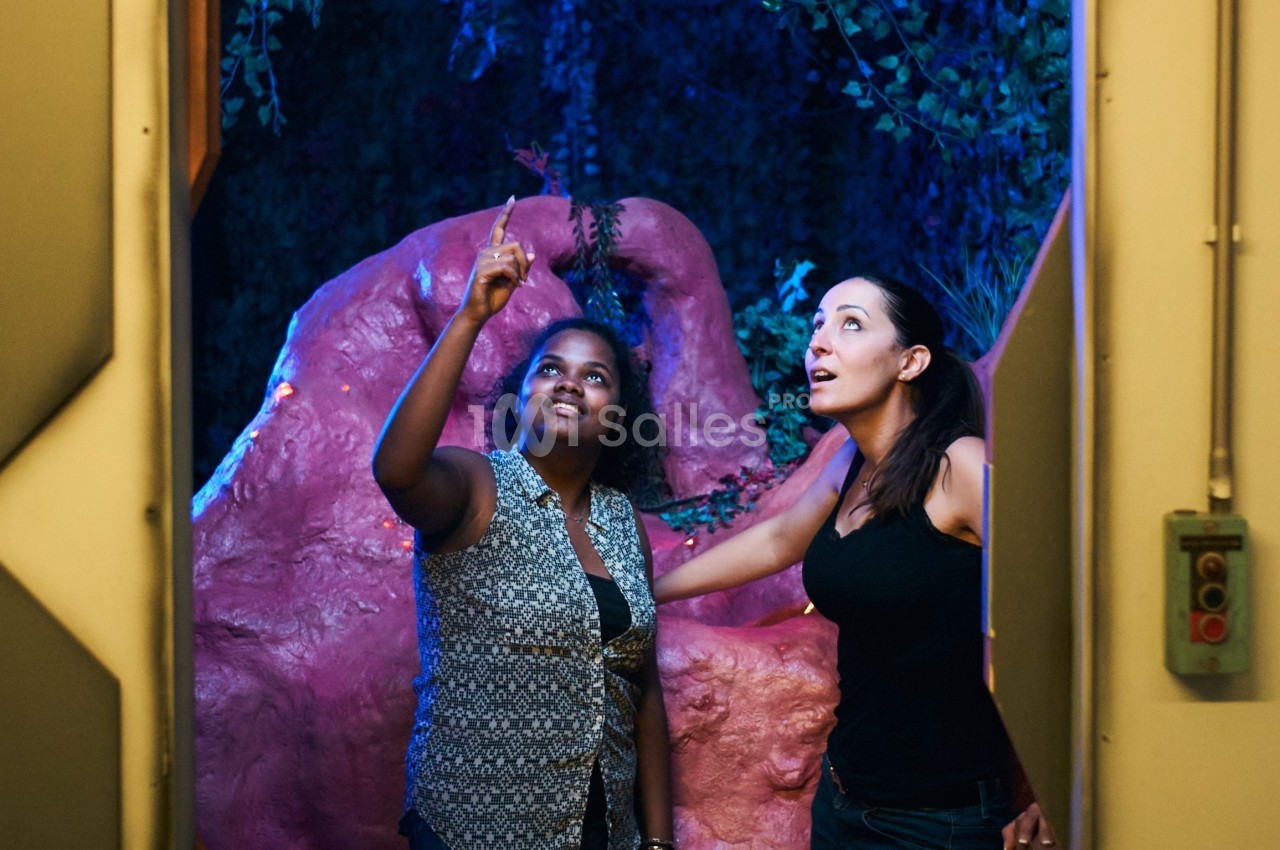 Deux femmes observent un élément lumineux dans un décor naturel avec des plantes et des rochers roses.