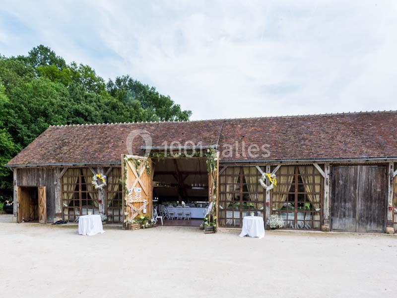 Grange en bois avec portes ouvertes, décorée pour un événement, entourée de verdure et d'une cour dégagée.