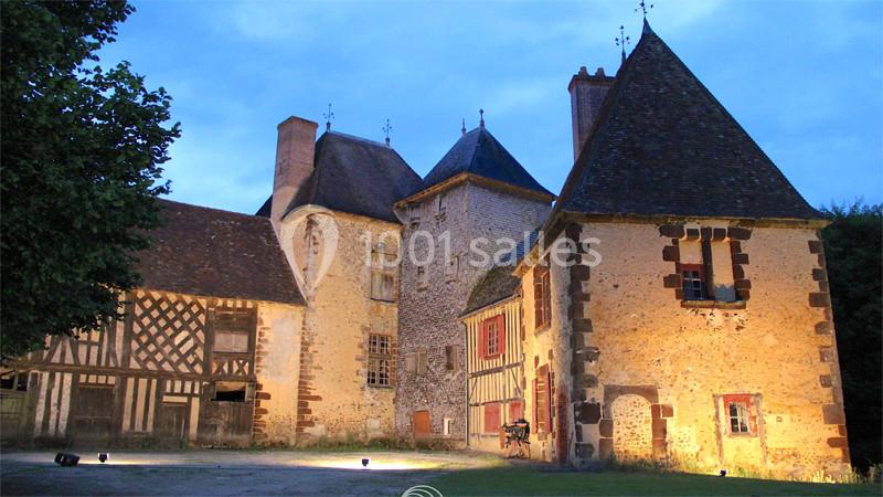 Façade éclairée d'un manoir médiéval en pierre et colombages, entouré de végétation, au crépuscule.