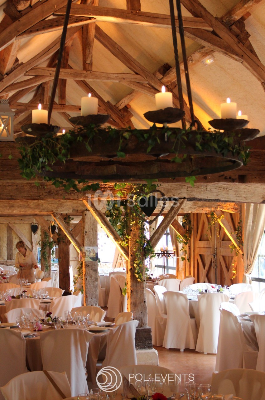 Salle de réception rustique avec poutres apparentes, tables dressées et lustre décoré de bougies et de feuillage.