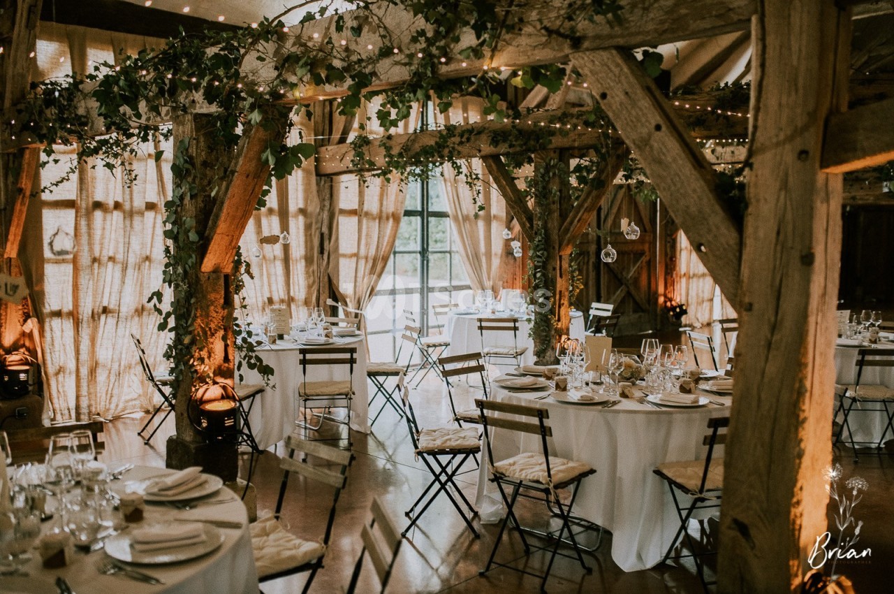Salle de réception rustique décorée de guirlandes lumineuses, de feuillages et de tables dressées pour un événement.
