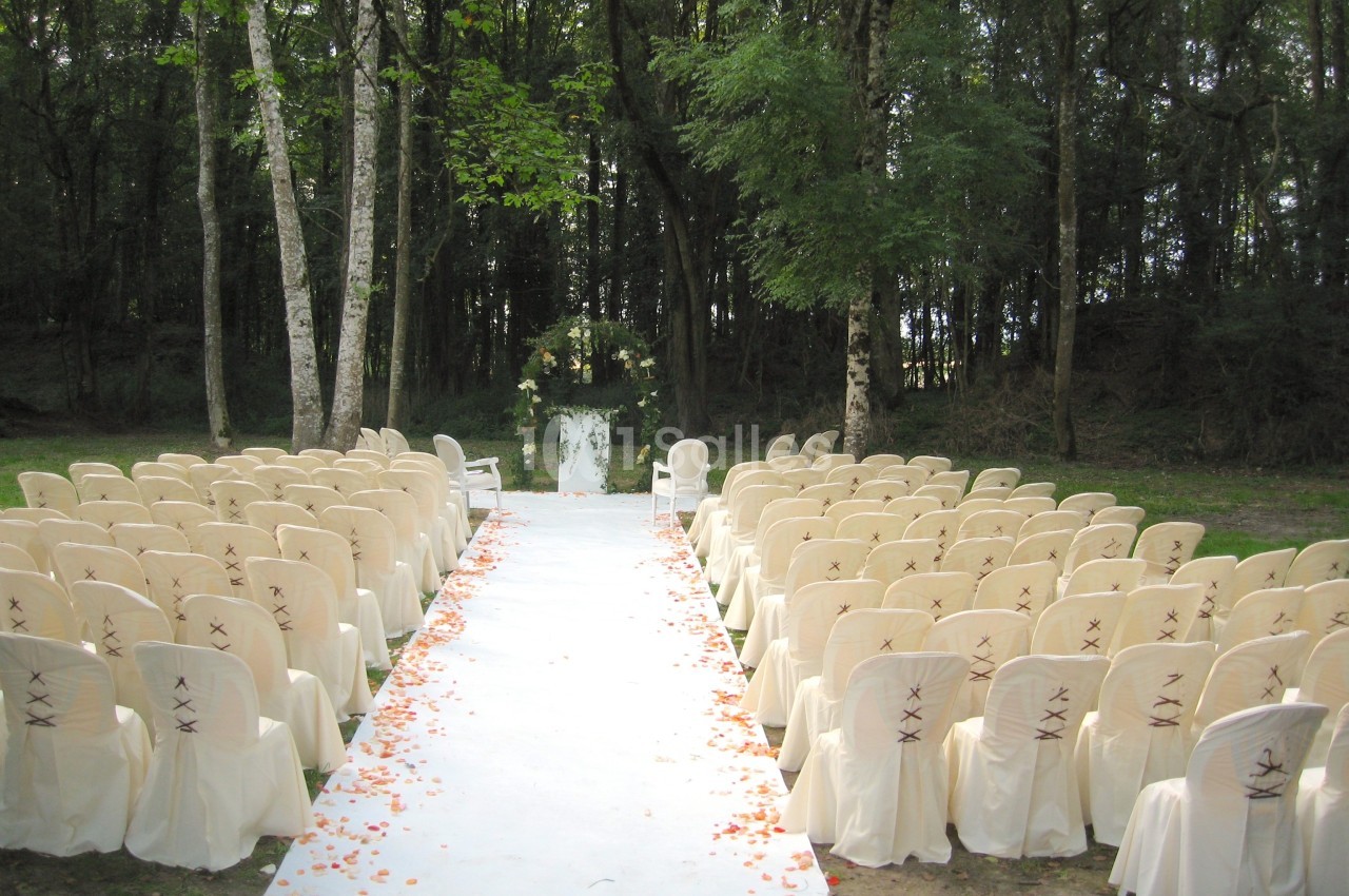Allée centrale bordée de chaises blanches décorées, menant à un autel floral en plein air dans une forêt.