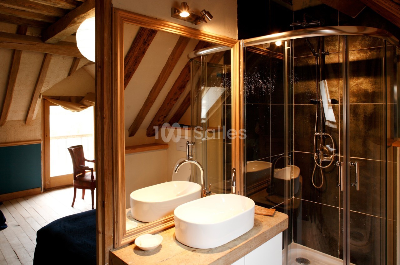 Salle de bain avec lavabo moderne, douche vitrée et miroir, dans une pièce aux poutres apparentes en bois.