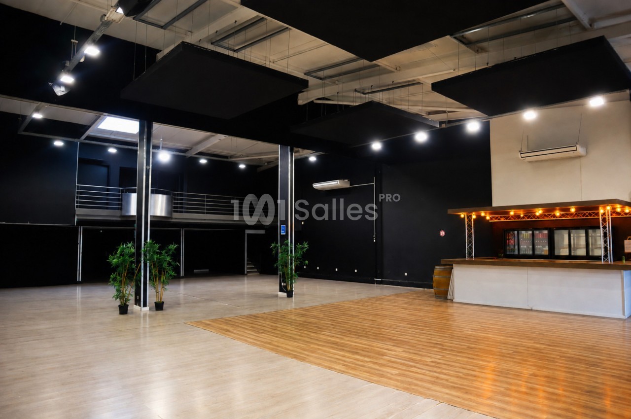 Salle spacieuse avec parquet, bar éclairé, plantes décoratives et mezzanine, éclairée par des spots au plafond.