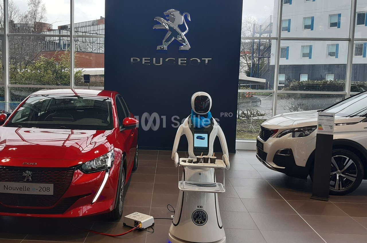 Un robot interactif exposé entre deux voitures Peugeot dans un showroom lumineux.