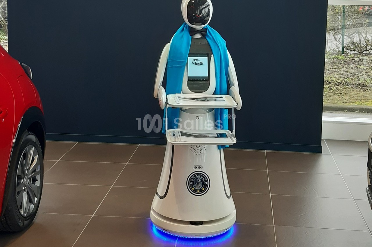 Robot blanc avec écran intégré, écharpe bleue et plateaux, exposé dans un espace intérieur avec des voitures en arrière-plan.