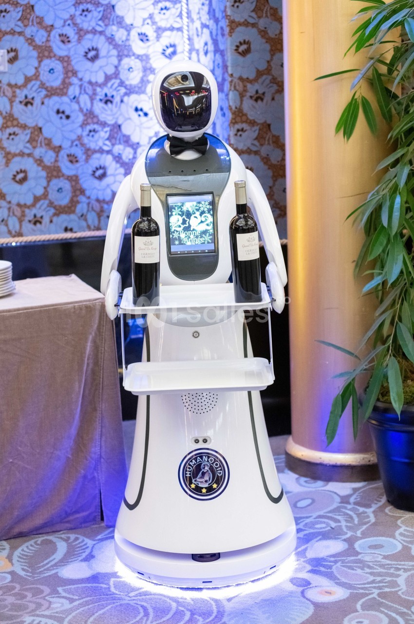 Robot blanc équipé d'un écran et portant deux bouteilles de vin sur un plateau, dans un environnement intérieur élégant.