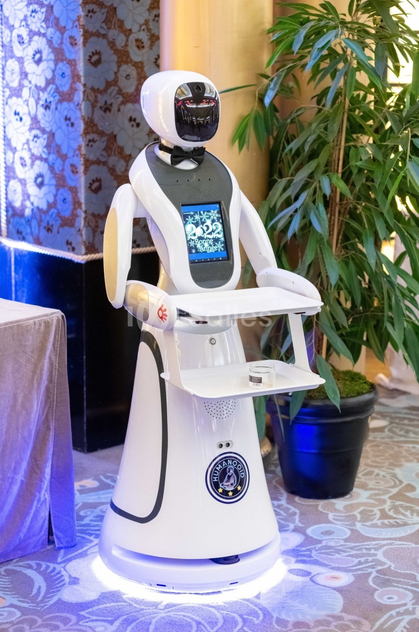 Robot humanoïde blanc équipé d'un écran et d'un plateau, présenté dans un environnement intérieur avec plantes et…