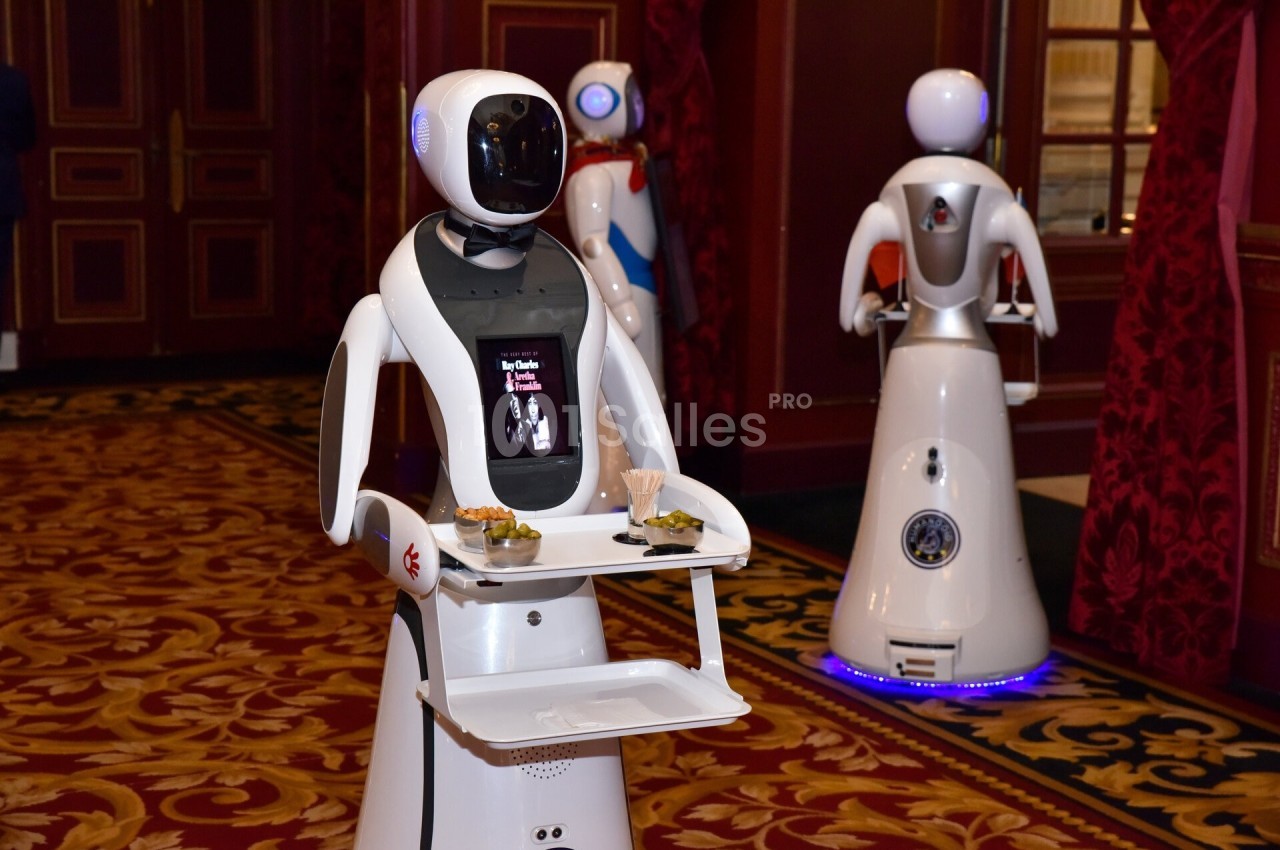 Des robots humanoïdes équipés de plateaux servent des amuse-bouches dans un intérieur élégant avec tapis rouge.