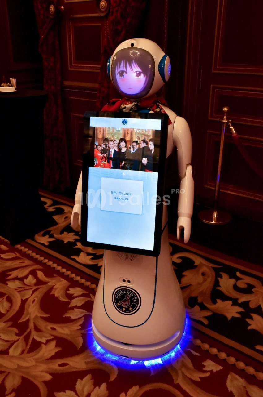 Robot humanoïde blanc avec écran affichant un visage animé, exposé dans une pièce ornée de rideaux rouges et tapis.