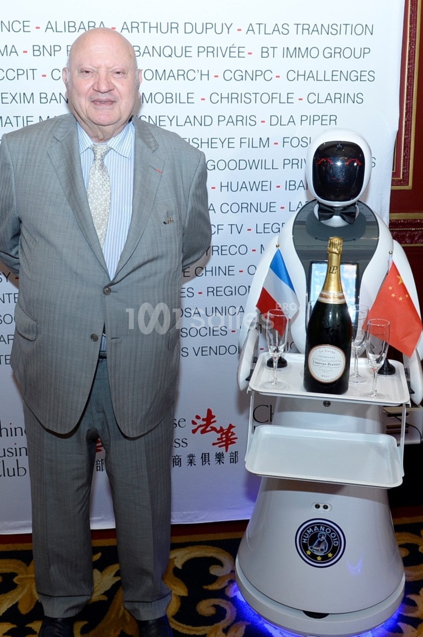 Un homme en costume pose à côté d'un robot portant une bouteille de champagne et des drapeaux français et chinois.