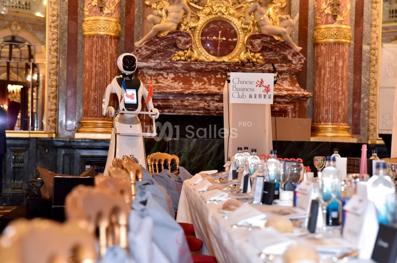Un robot humanoïde présenté dans une salle ornée, devant une table dressée pour un événement formel.