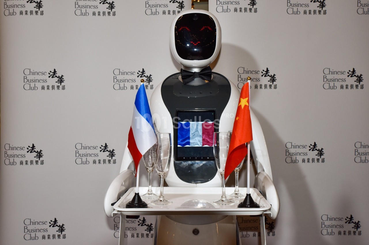 Un robot humanoïde portant un plateau avec des drapeaux français et chinois, devant un fond marqué ’Chinese Business Club’.