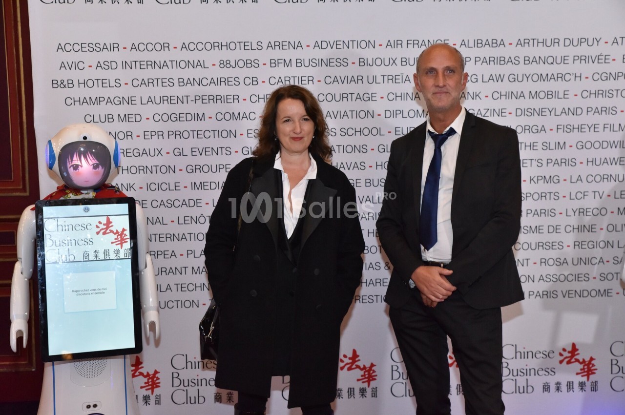 Un homme et une femme posent debout à côté d'un robot avec un écran tactile, devant un mur de logos.