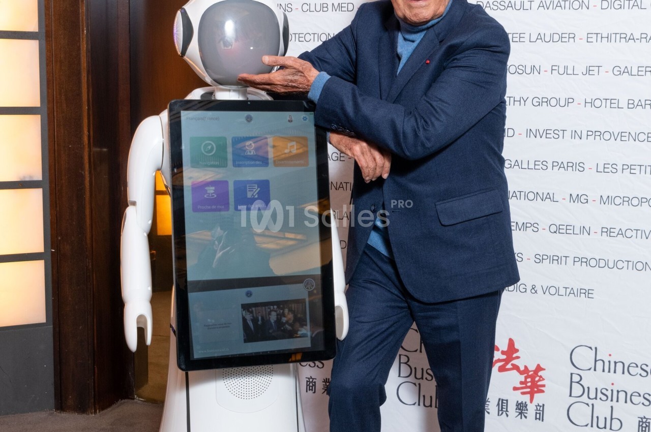 Un homme souriant pose à côté d'un robot interactif équipé d'un écran tactile, dans un cadre professionnel.