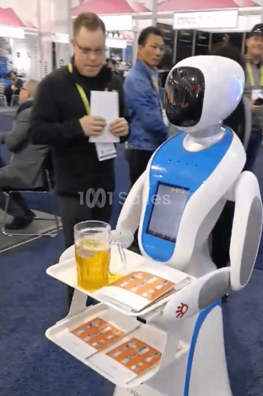 Un robot serveur blanc et bleu portant une chope de bière et des plateaux, entouré de personnes dans un salon professionnel.