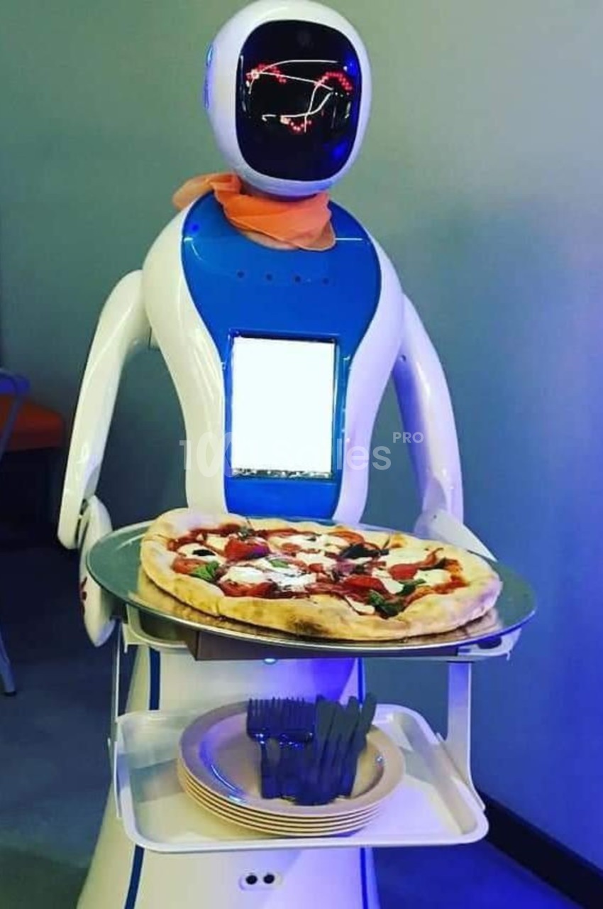 Robot humanoïde bleu et blanc servant une pizza sur un plateau, avec des assiettes et des couverts en dessous.
