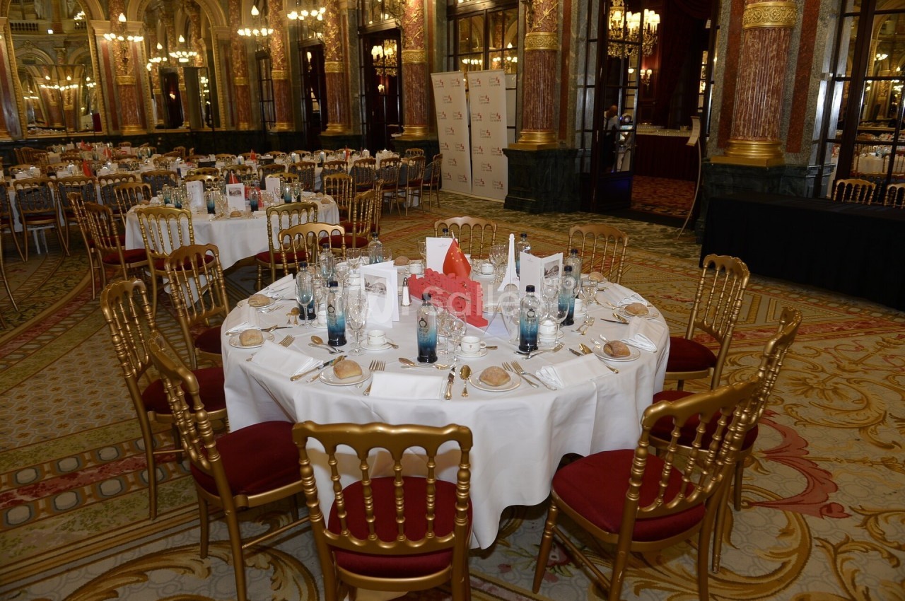 Salle de réception élégante avec tables rondes dressées, nappes blanches, chaises dorées et décorations raffinées.