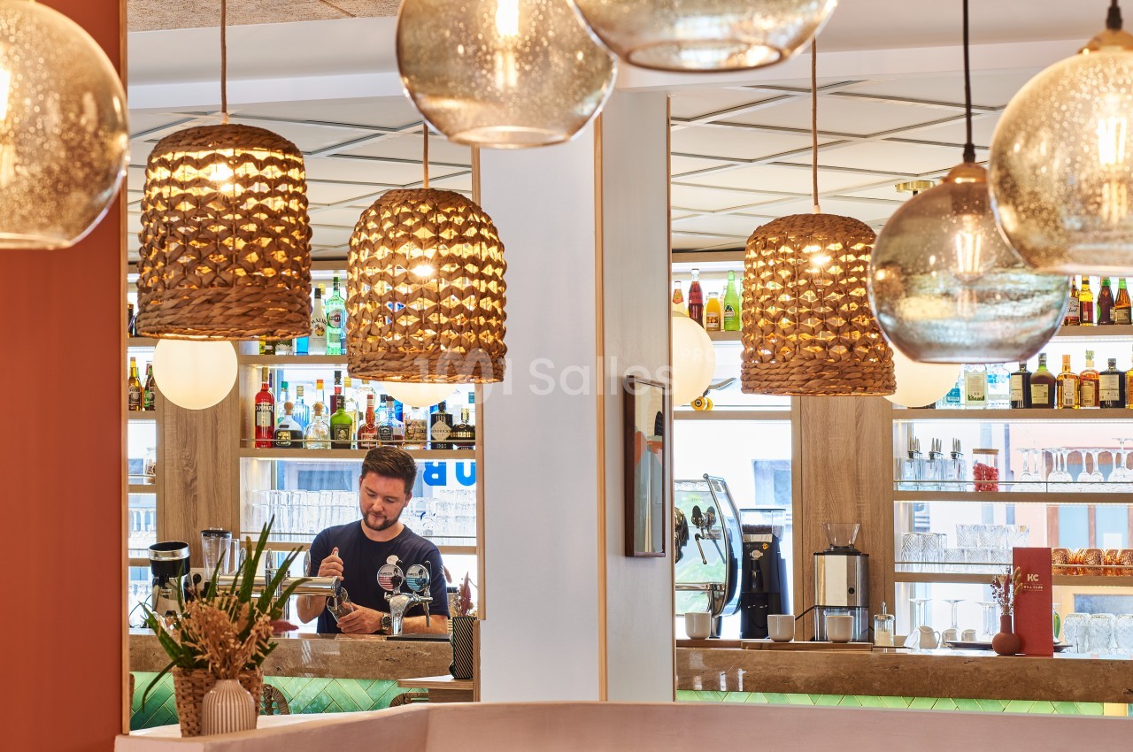 Un barman prépare une boisson derrière un comptoir, entouré de bouteilles et de suspensions lumineuses décoratives.