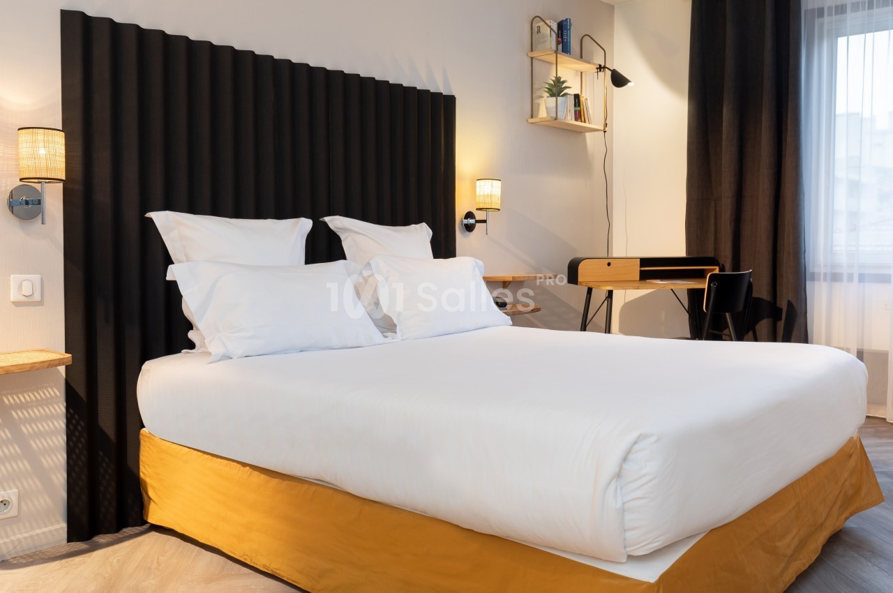 Lit double avec draps blancs, tête de lit noire, bureau en bois avec chaise et étagères murales dans une chambre lumineuse.