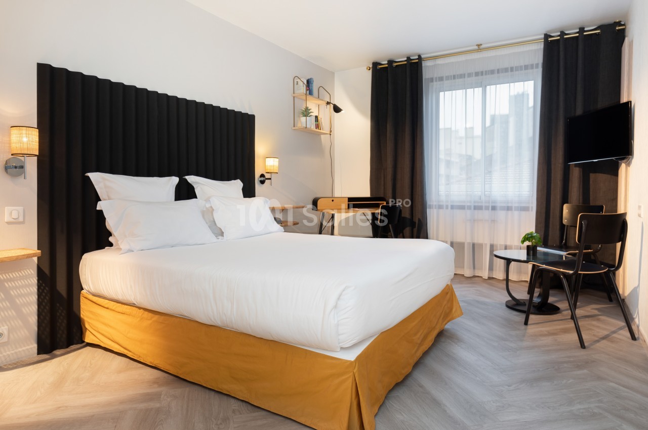 Chambre lumineuse avec lit double, bureau, télévision murale, rideaux sombres et fenêtre donnant sur l'extérieur.