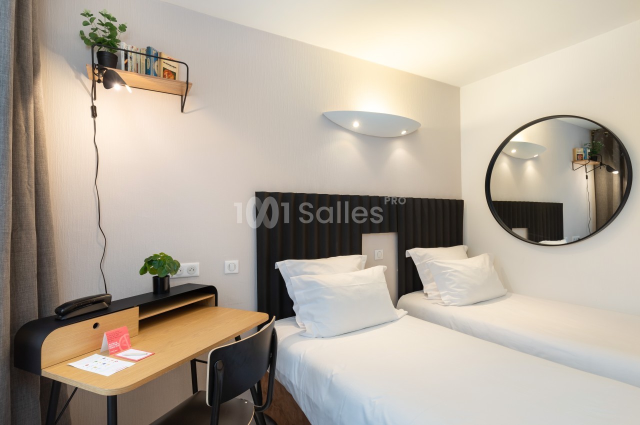 Chambre avec deux lits simples, bureau en bois, chaise noire, étagère murale et miroir rond sur un mur clair.