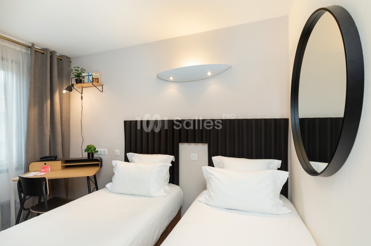Deux lits simples avec tête de lit noire, bureau en bois, chaise noire, miroir rond et lumière naturelle.