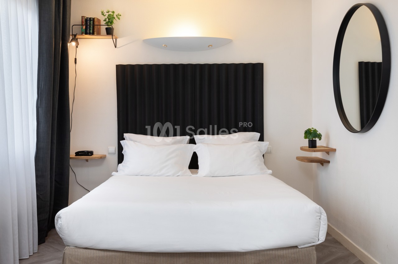 Lit double avec draps blancs dans une chambre moderne, éclairée par une applique murale, avec étagères et miroir rond.
