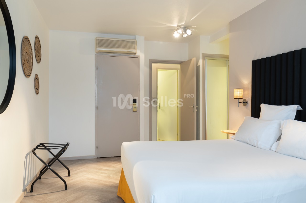 Chambre lumineuse avec lit double, murs blancs, décoration murale, miroir rond et porte ouverte sur un couloir.
